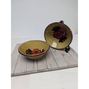 May include: Two ceramic bowls with a floral design. The bowls are a light yellow color with a dark brown rim. The floral design is red and black. One bowl is upright, and the other is tilted on a black stand.