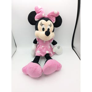 Op de afbeelding: Een roze jurk met witte stippen en een zwarte strik op het hoofd. Het pluche speelgoed is een afbeelding van Minnie Mouse.
