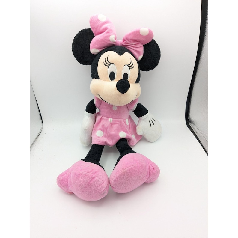 Op de afbeelding: Een roze jurk met witte stippen en een zwarte strik op het hoofd. Het pluche speelgoed is een afbeelding van Minnie Mouse.