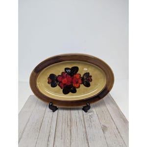 May include: Oval ceramic serving platter with a brown rim and a beige interior. The center features a floral design in red, black, and brown. The platter is displayed on a black stand.