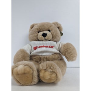 May include: A brown teddy bear wearing a white sweater with the red text "Lambert Kay" on the front.