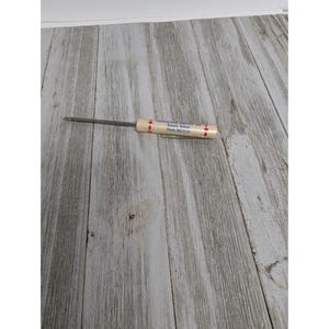 May include: A vintage screwdriver with a cream-colored handle and a metal shaft. The handle has red arrow designs and text that reads "Russell, Kansas Phone 483-3116". The tool is designed for turning screws and is likely used for repairs or construction.