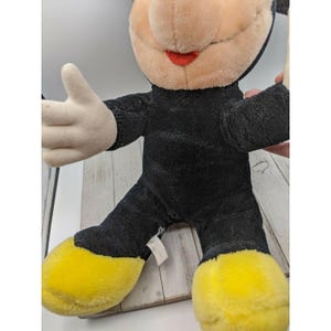 May include: A black and yellow plush Mickey Mouse toy with a red mouth. The toy is sitting on a wooden surface.