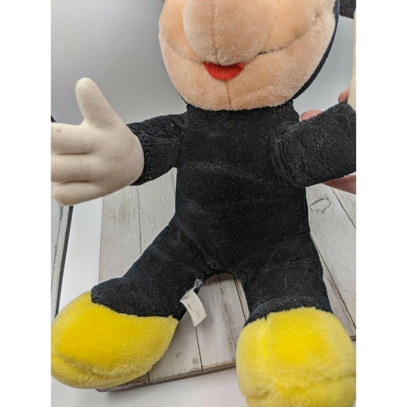 May include: A black and yellow plush Mickey Mouse toy with a red mouth. The toy is sitting on a wooden surface.
