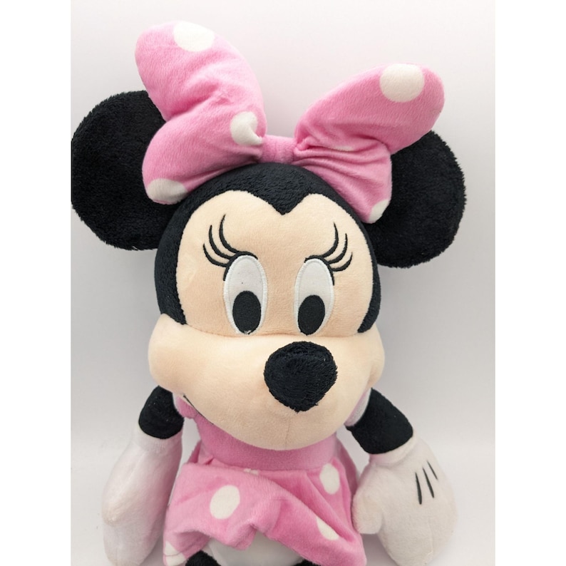 Op de afbeelding: Een roze en wit gestippeld pluche speelgoed van Minnie Mouse. Ze heeft een grote zwarte strik en grote zwarte oren.