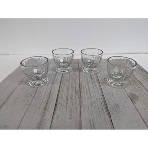 May include: Four clear glass shot glasses with a textured, dimpled design. The glasses have a rounded bowl shape and a sturdy base. They are arranged on a light gray wooden surface.