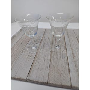 May include: Two clear, stemmed wine glasses with etched floral designs. The glasses have a flared bowl and a slender stem, resting on a circular base. The glasses are displayed on a light gray wooden surface.