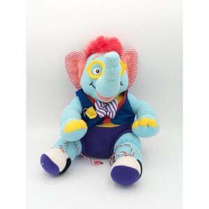 May include: A blue plush elephant toy wearing a blue waistcoat, purple trousers, and a bow tie. The elephant has red hair, yellow spectacles, and a red and white striped ear.