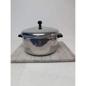 May include: A stainless steel cooking pot with a lid and black handles. The pot is sitting on a gray wooden surface. The lid has a black knob. The pot is round and has a shiny, reflective surface.