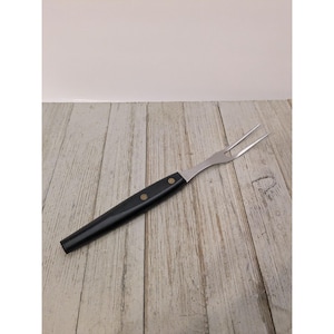 Puede incluir: Un tenedor de trinchar de dos puntas con una cabeza larga y delgada de metal plateado y un mango negro liso y sólido. El mango está fijado a la espiga de metal con dos pequeños remaches circulares de color latón visibles en el lateral.