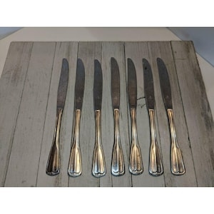 May include: A collection of seven silver butter knives with decorative handles. The blades are a dull silver, and the handles feature a scalloped design. The knives are displayed on a light grey wooden surface.