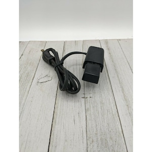 Puede incluir: Un adaptador de corriente negro con un cable y un enchufe. El adaptador es rectangular y tiene un pequeño botón negro en el lateral.