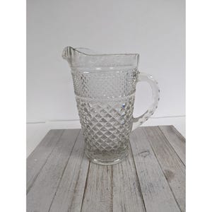 May include: Clear glass pitcher with a diamond pattern design. The pitcher has a handle and a spout for pouring. The pitcher is sitting on a wooden surface.