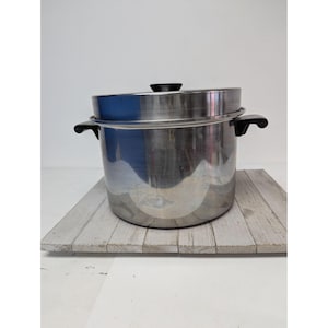 May include: A stainless steel steamer pot with a black handle and lid. The pot has a shiny, reflective surface and is sitting on a wooden surface. The steamer pot is designed for cooking vegetables and other foods.