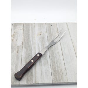May include: A stainless steel carving fork with a dark brown wooden handle. The fork has two tines and is designed for handling and serving roasted meats. The handle is secured with two silver rivets.