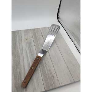 May include: A stainless steel spatula with a slotted head and a wooden handle. The handle is a dark brown color and is attached to the metal head with a metal rivet. The spatula is designed for grilling or cooking.