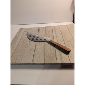 Puede incluir: Un cuchillo de cocina con mango de madera y hoja de metal plateado curvada. La hoja presenta una serie de ranuras rectangulares horizontales a lo largo de su longitud, diseñadas para cortar. El mango está asegurado con dos remaches.