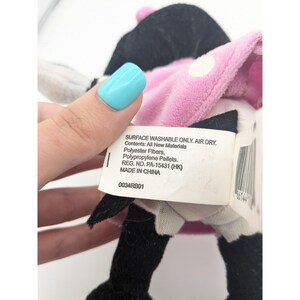 Puede incluir: Un juguete de peluche negro y blanco con detalles rosas. El juguete tiene un lazo negro y una cara blanca con ojos negros. El juguete lleva un vestido rosa con lunares blancos. El juguete tiene una etiqueta que dice "SURFACE WASHABLE ONLY. AIR DRY. Contents: All New Materials Polyester Fibers, Polypropylene Pellets. REG. NO. PA-15431 (HK) MADE IN CHINA 0034RB01".