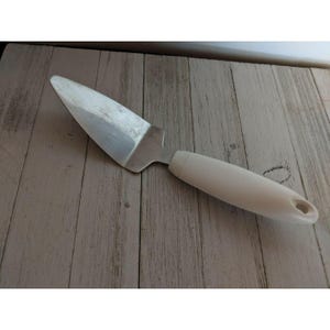 Puede incluir: Un servidor de pastel de acero inoxidable con un mango blanco. El servidor tiene una hoja triangular y un mango redondeado con un agujero en el extremo. Diseñado para servir pasteles y tartas.