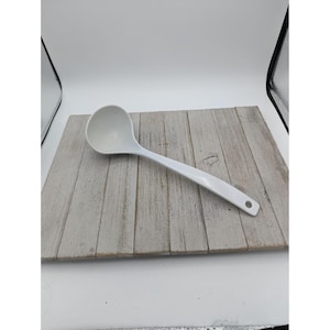 May include: A white ladle with a long handle and a round bowl. The ladle is made of a solid, opaque material and has a small hole at the end of the handle for hanging. The ladle is resting on a light gray wooden surface.