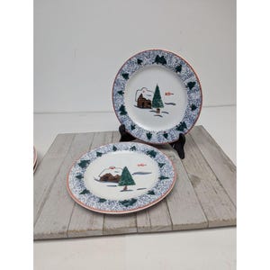 May include: Two white ceramic plates with a winter scene. Each plate features a cabin, a green tree, and a red and blue border. The plates are displayed on a wooden surface.