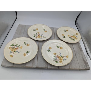 May include: Four cream-coloured plates with a floral design of yellow and blue flowers and green leaves. Each plate has a gold rim. The plates are arranged on a weathered wooden surface.