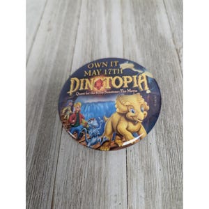 May include: A round button with a blue background and a yellow dinosaur on it. The button says "Own it May 17th Dinotopia Quest for the Ruby Sunstone: The Movie".