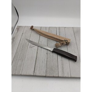 May include: A stainless steel knife with a dark brown handle and a tan leather sheath. The knife is laying on a light gray wooden surface. The sheath is open and next to the knife.