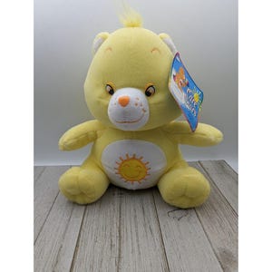 May include: Yellow plush Care Bear with a smiling sun on its belly. The bear has a white face with black eyes and a pink nose. The Care Bears logo is on the tag.