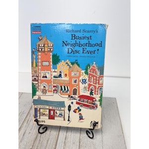 May include: A colorful CD case featuring the title "Richard Scarry's Busiest Neighborhood Disc Ever!" The cover depicts a vibrant cartoon neighborhood with buildings like a City Hall, bakery, and post office. A red bus and characters are also visible.