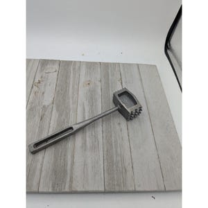 May include: A silver meat tenderizer mallet with a rectangular head and a long handle. The head has a textured surface on one side and a flat surface on the other. The handle has a rectangular hole.