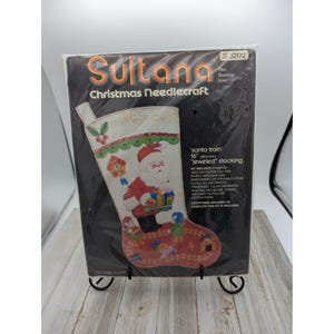 May include: A needlepoint kit for a Christmas stocking featuring a Santa Claus with a train and the text 'santa train' 16" (40.6 cm) 'jeweled' stocking. The kit includes stamped 100% polyester felt for sample applique and embroidery, six strand cotton floss, all decorative trimmings, colour separated sequins and beads, needle and easy to follow instructions.