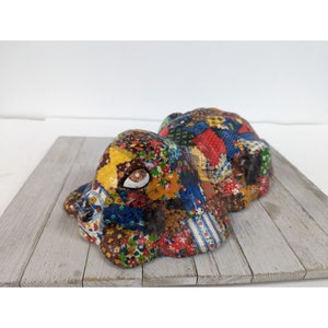 Könnte beinhalten: Eine Keramikskulptur eines schlafenden Hundes, bedeckt mit einem Patchworkmuster aus bunten Stoffen. Das Auge des Hundes ist mit einer weißen Pupille und einer braunen Iris bemalt.