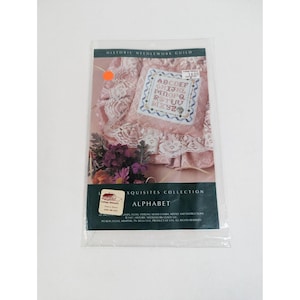 May include: A cross-stitch kit featuring an alphabet design. The kit includes a pattern with the letters A through Z, displayed on a pink fabric with floral accents. The packaging reads "Historic Needlework Guild" and "Alphabet."
