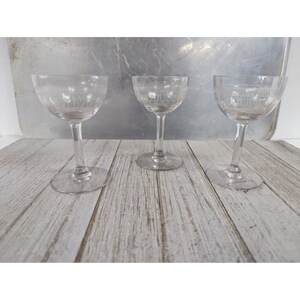 May include: Three clear glass stemmed drinking glasses with etched designs. The glasses are arranged in a row on a light wood surface.