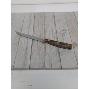 May include: A single steak knife with a stainless steel blade and a wooden handle. The handle is a dark brown color with brass rivets. The knife is resting on a light gray wooden surface.