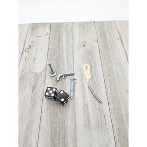 May include: A collection of miniature game pieces, including two black dice with white dots, a silver toy gun, a wrench, a rope, and a silver screw. The items are arranged on a light wood-grain surface.