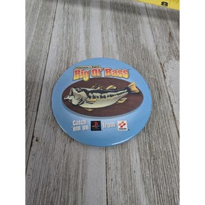 May include: A blue and white round button with a picture of a bass fish and the text "Fisherman's Bait 2 Big or Bass Catch 'em on PlayStation from Konami".