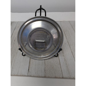 Puede incluir: Un plato redondo de metal plateado con asa, exhibido en un soporte de metal negro. El plato tiene un centro ligeramente cóncavo y un borde elevado. El asa es rectangular y está fijada a la superficie del plato.