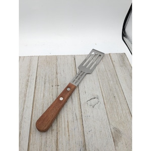 May include: A stainless steel spatula with a wooden handle. The spatula has a slotted design and the text "Grandma's Kitchen" is engraved on the handle.