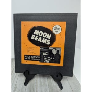 May include: Vintage vinyl record album titled "Moon Beams" by Paul Carson at the NBC Organ. The album cover is orange with black text and a photo of Carson playing the organ. The album is in a black frame.