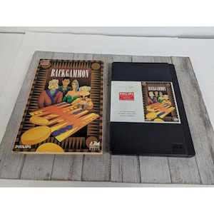 May include: A vintage Philips Interactive Backgammon game for the Magnavox Odyssey 2. The box is black with a white label that says "Backgammon" and features a cartoon image of four people playing the game. The game manual is yellow with a cartoon image of a backgammon board and four people playing the game. The manual cover says "Backgammon" in large letters.