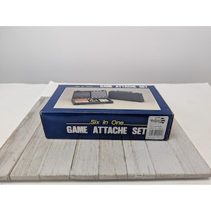 May include: A blue box set labeled "Six In One GAME ATTACHE SET" with a picture of the game pieces. The box is on a white and gray wooden surface. The set includes a checkerboard and other game components.
