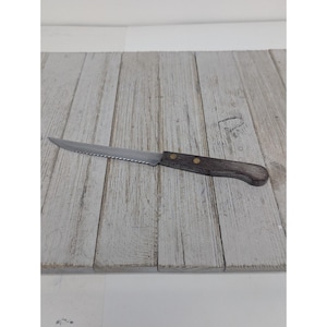 May include: A single steak knife with a serrated stainless steel blade and a dark wooden handle. The handle is secured with two brass rivets. The knife is designed for cutting meat.