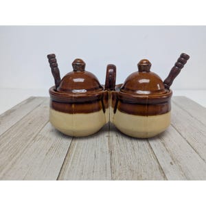 Op de afbeelding: Een tweedelige keramische condimentenserver met een handvat, elke sectie bekroond met een deksel en een houten lepel. De server is bruin en crèmekleurig, met een glanzende afwerking. De lepels zijn donkerbruin.