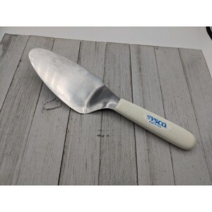 May include: A white handled cake server with a stainless steel blade. The handle has the text "Sysco Made in U.S.A." printed in blue.