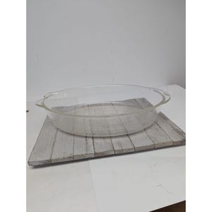May include: A clear, oval-shaped glass baking dish with handles. The dish is sitting on a gray and white wooden plank. The dish is empty and appears to be unused. Ideal for baking casseroles, desserts, or other dishes.