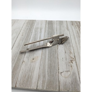 Puede incluir: Un abrelatas vintage de color plateado, con un mango largo y una rueda de corte accionada por engranajes. El abrelatas está diseñado para perforar y cortar las tapas de las latas. La herramienta es de metal y parece bien usada.