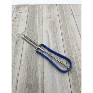 May include: A vintage vegetable peeler with a stainless steel blade and a blue handle. The peeler is designed with a loop handle for easy grip and control. The tool is ideal for peeling fruits and vegetables.