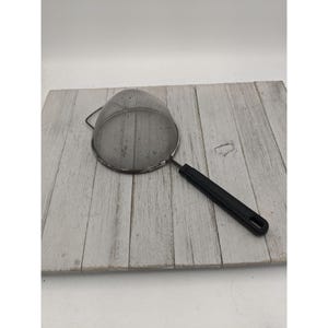 May include: A stainless steel mesh strainer with a black plastic handle. The strainer is round with a fine mesh and a metal rim. The handle has a hole for hanging. The strainer is designed for straining liquids or sifting dry ingredients.